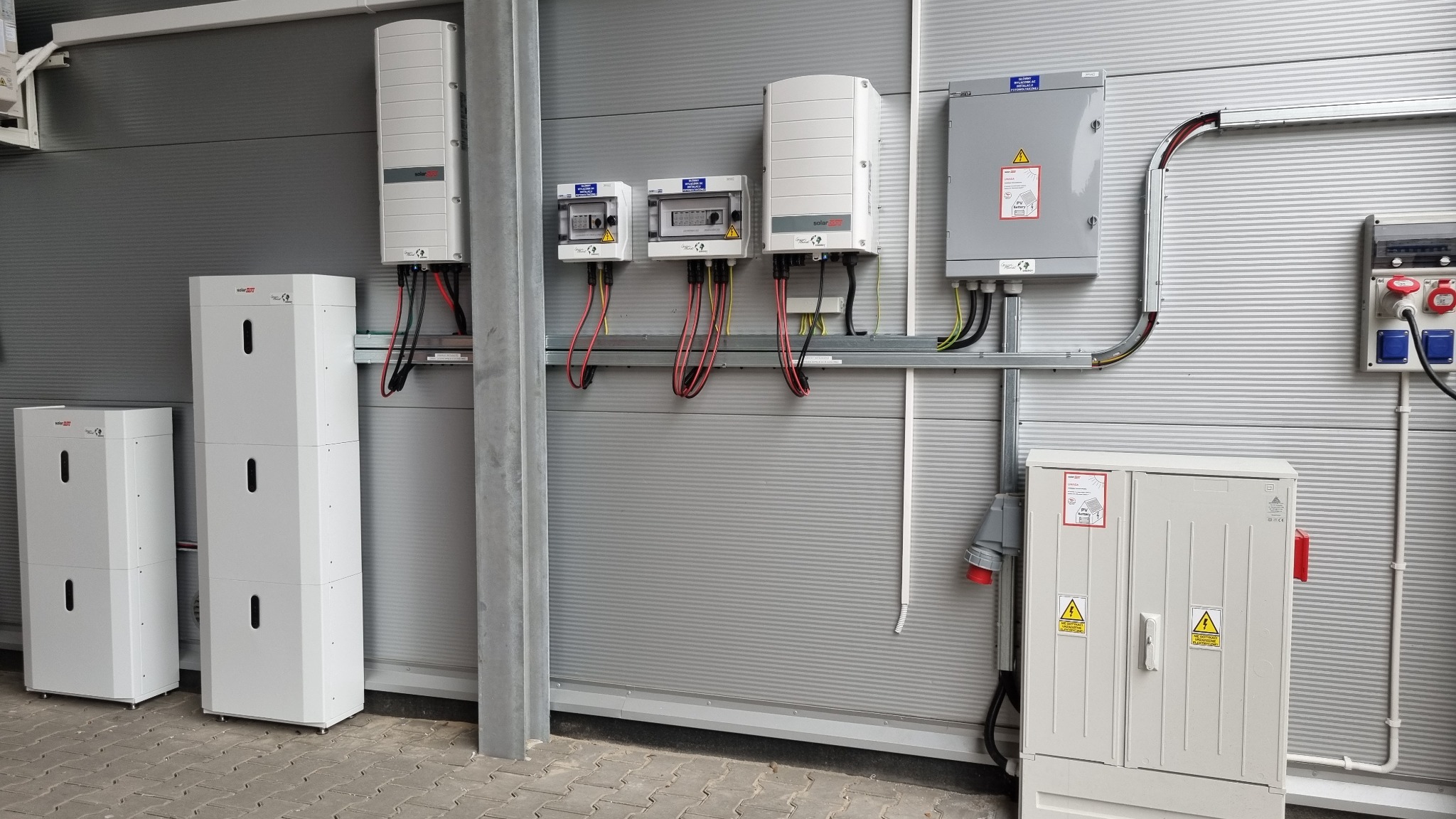 Kompletna instalacja z magazynem energii: białe moduły bateryjne, inwertery i rozdzielnice na szarej ścianie z blachy trapezowej. Widoczne kable i oznaczenia bezpieczeństwa.