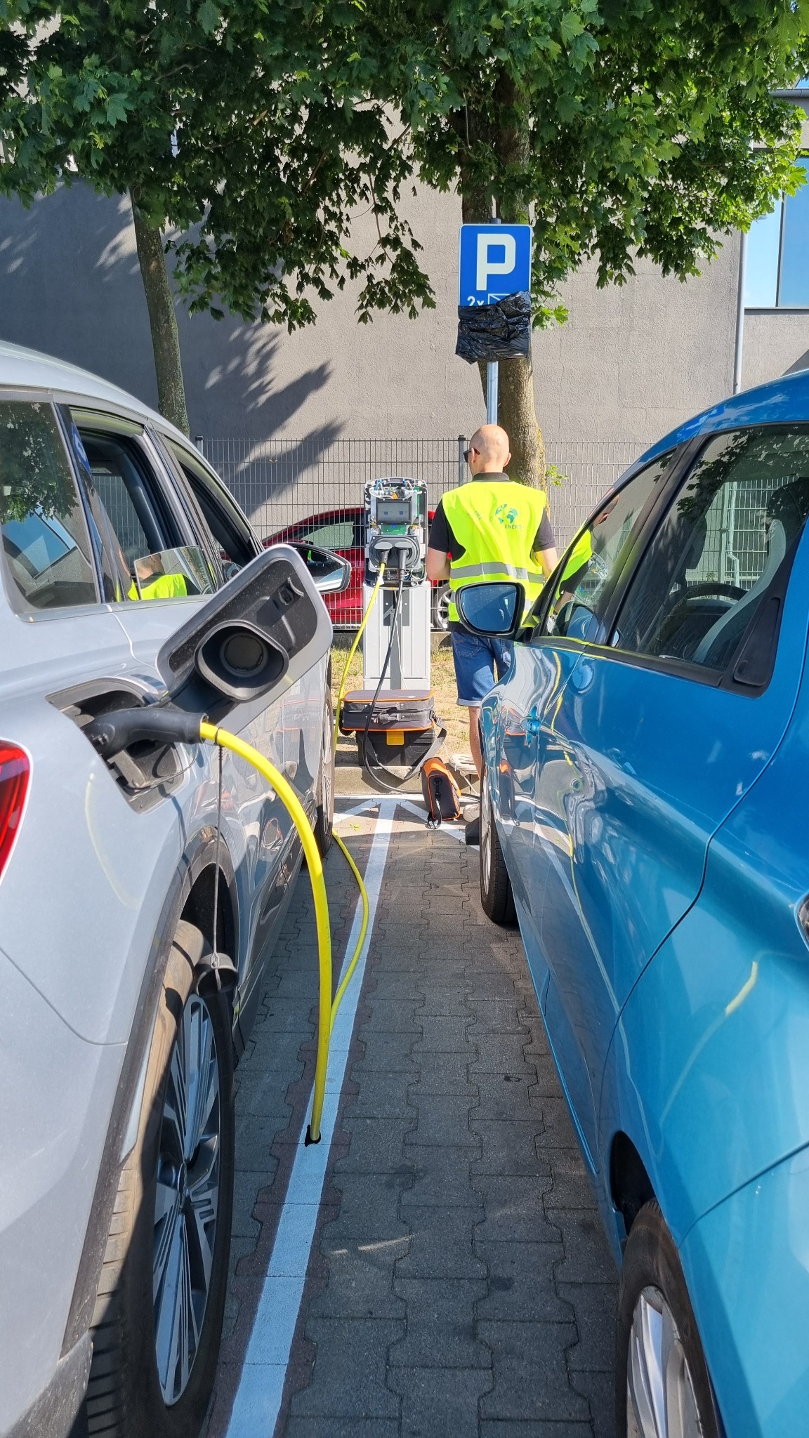 Dwa samochody elektryczne ładowane na stacji z żółtym kablem, pracownik w kamizelce przy stacji, znak parkingu z workiem, drzewo w tle, kostka brukowa.