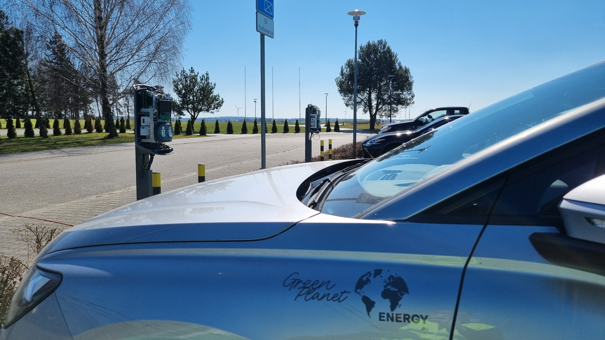 Samochód elektryczny z logo 'Green Planet Energy' na tle stacji ładowania EV i krajobrazu z turbinami wiatrowymi w oddali, w słoneczny dzień.