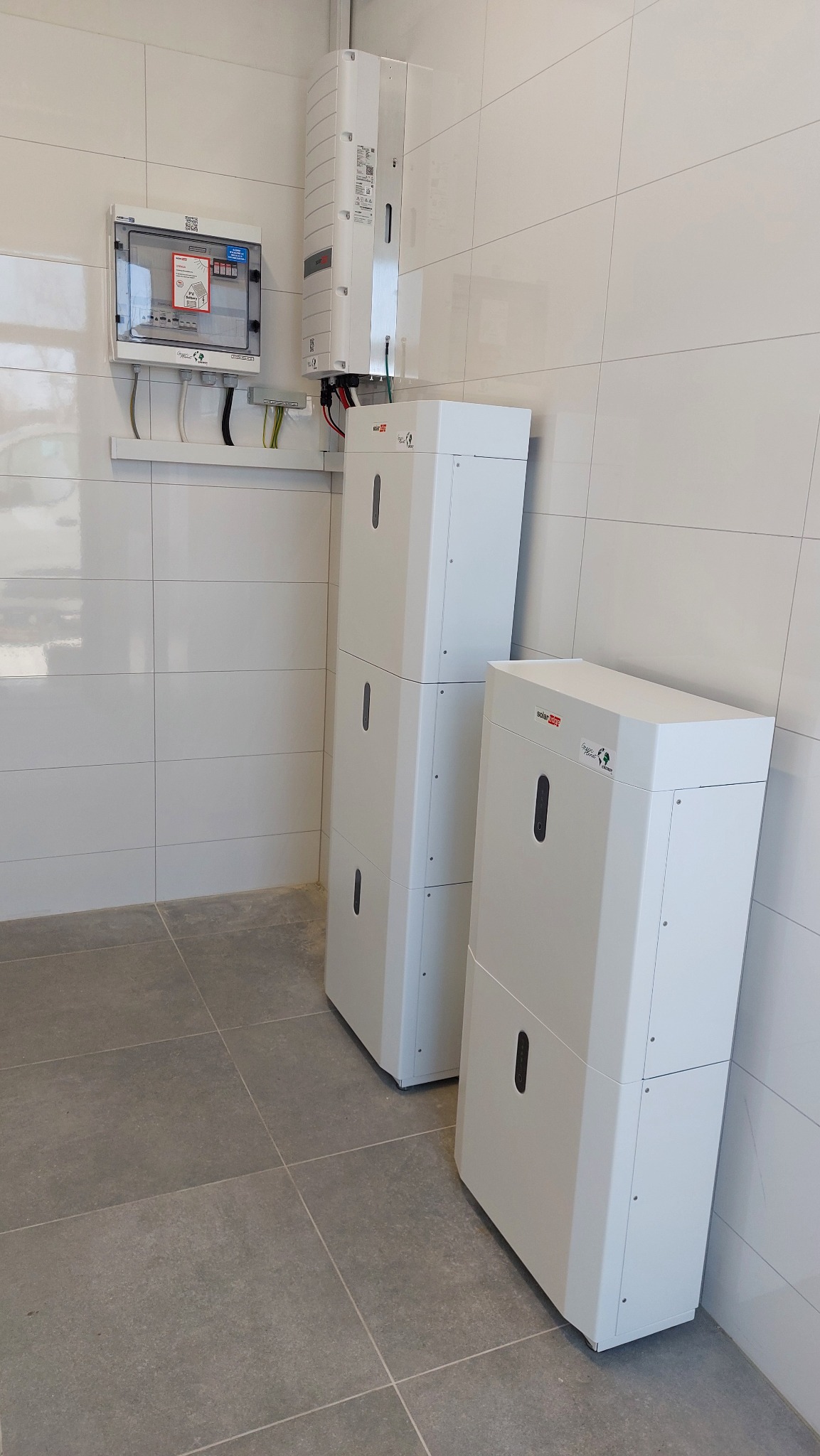 Dwa białe moduły magazynu energii SolarEdge w Gdyni, ustawione jeden obok drugiego, z widoczną skrzynką elektryczną i inwerterem na ścianie z białych płytek.