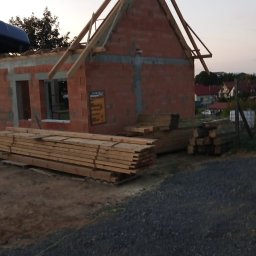 Mateusz Graf - Budowa domu jednorodzinnego w trakcie realizacji: ściany z czerwonej cegły, drewniana konstrukcja dachu, deski budowlane obok. Widok na okoliczne domy.