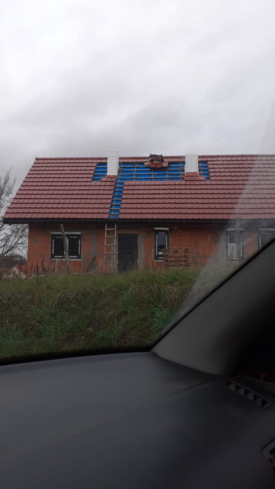 Budowa domu z czerwonej cegły, widok zza szyby samochodu. Częściowo położony dach z czerwoną dachówką, widoczne kominy i drabina oparta o ścianę. Pochmurne niebo.