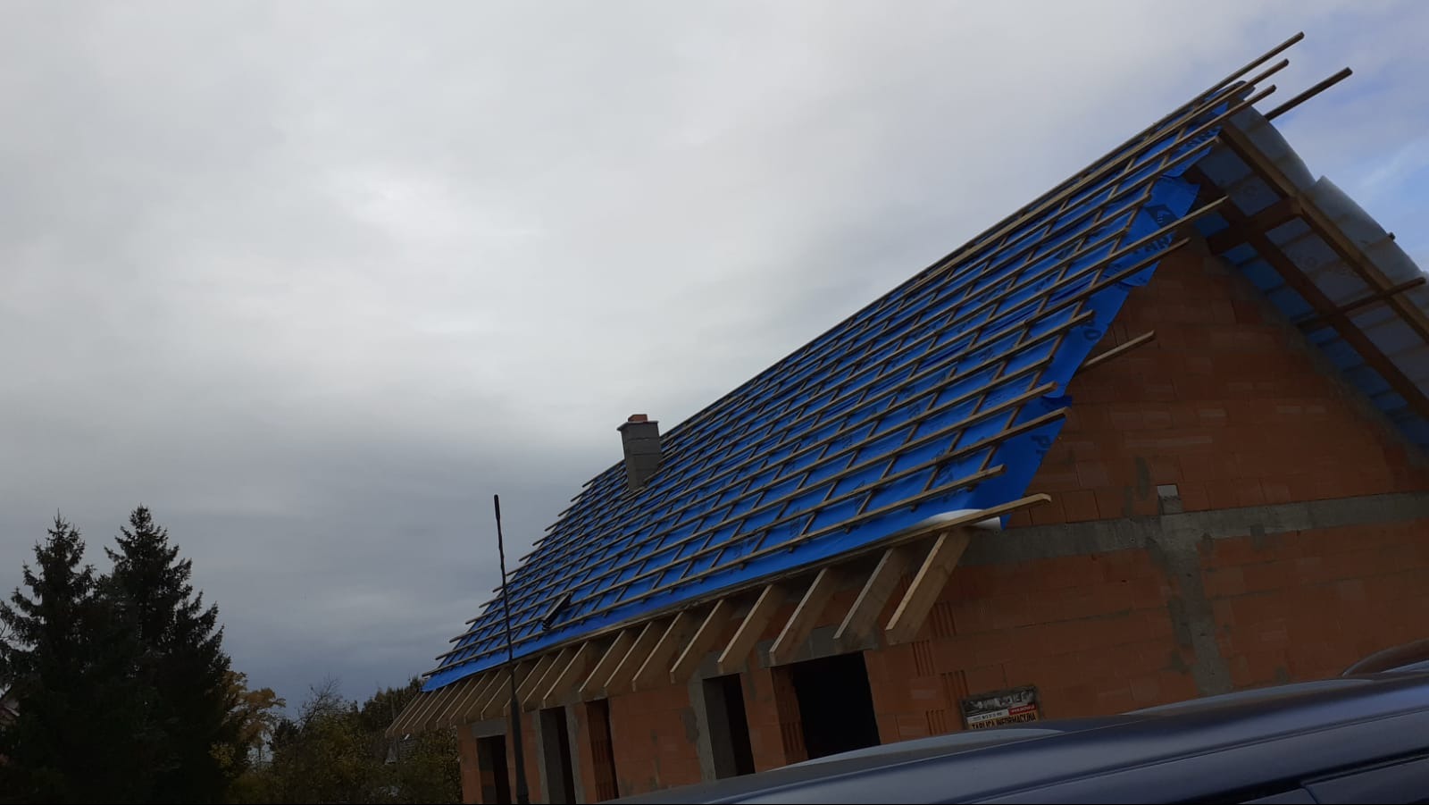 Budowa domu w trakcie: ceglany budynek z niebieską membraną dachową i drewnianą konstrukcją więźby, widok z dołu na tle pochmurnego nieba.