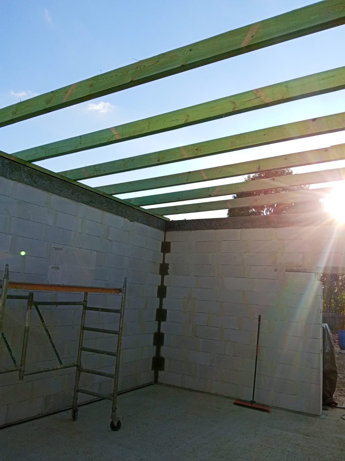 Wnętrze surowego budynku z bloczków, z widocznym drewnianym szkieletem dachu i metalowym rusztowaniem. Słońce wpada przez otwór w dachu.