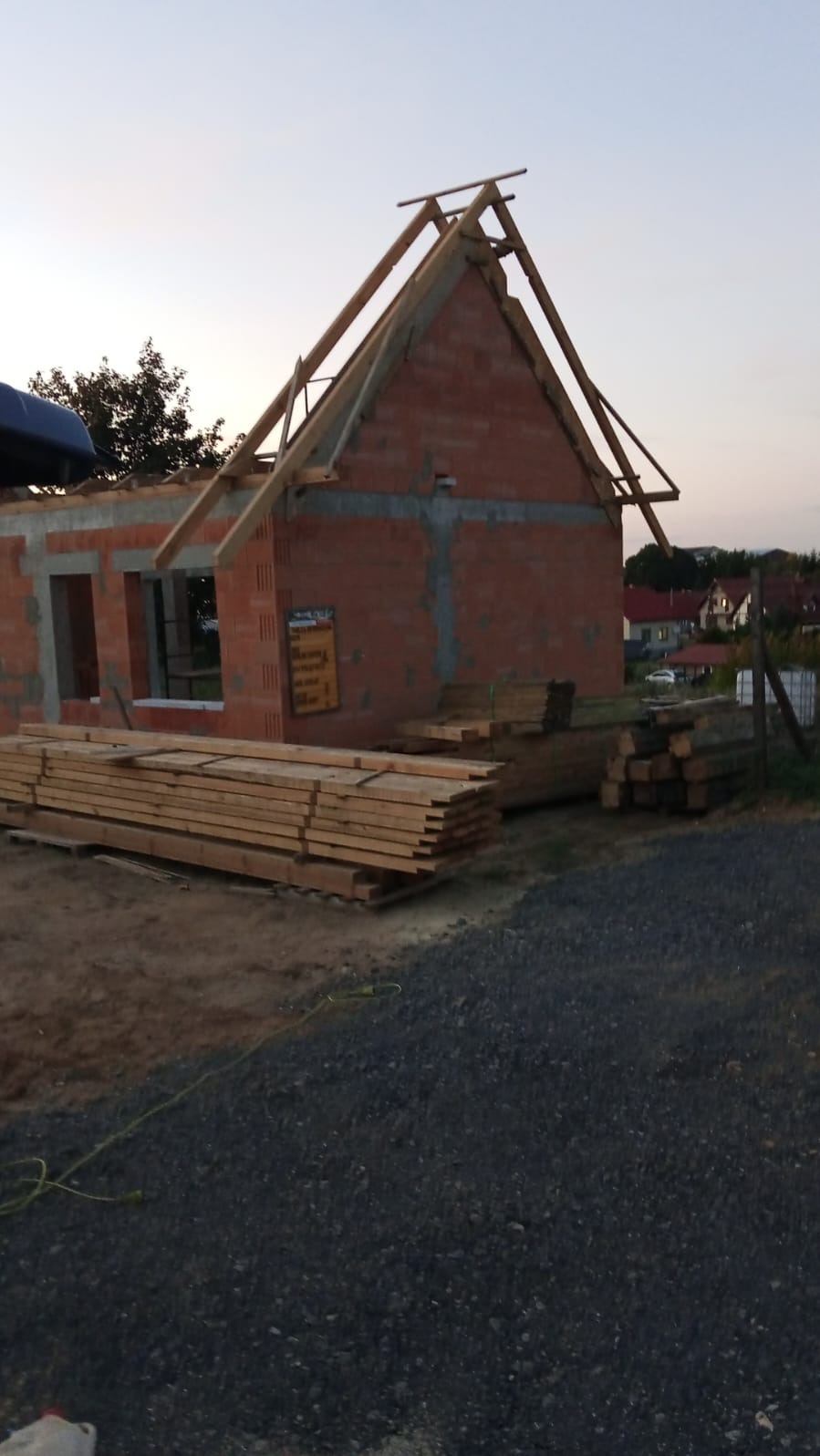 Budowa domu jednorodzinnego w trakcie realizacji: ściany z czerwonej cegły, drewniana konstrukcja dachu, deski budowlane obok. Widok na okoliczne domy.