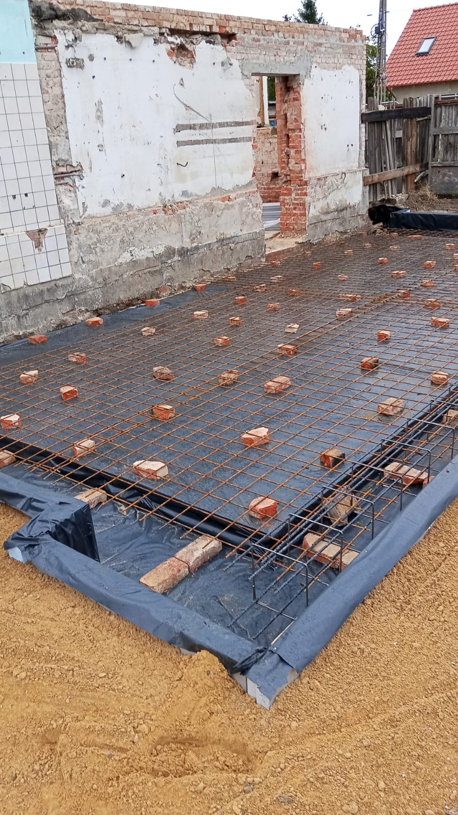 Zbrojenie fundamentów na czarnej folii, cegły dystansowe, widoczne fragmenty starego muru. Przygotowanie do wylania betonu, etap prac budowlanych.