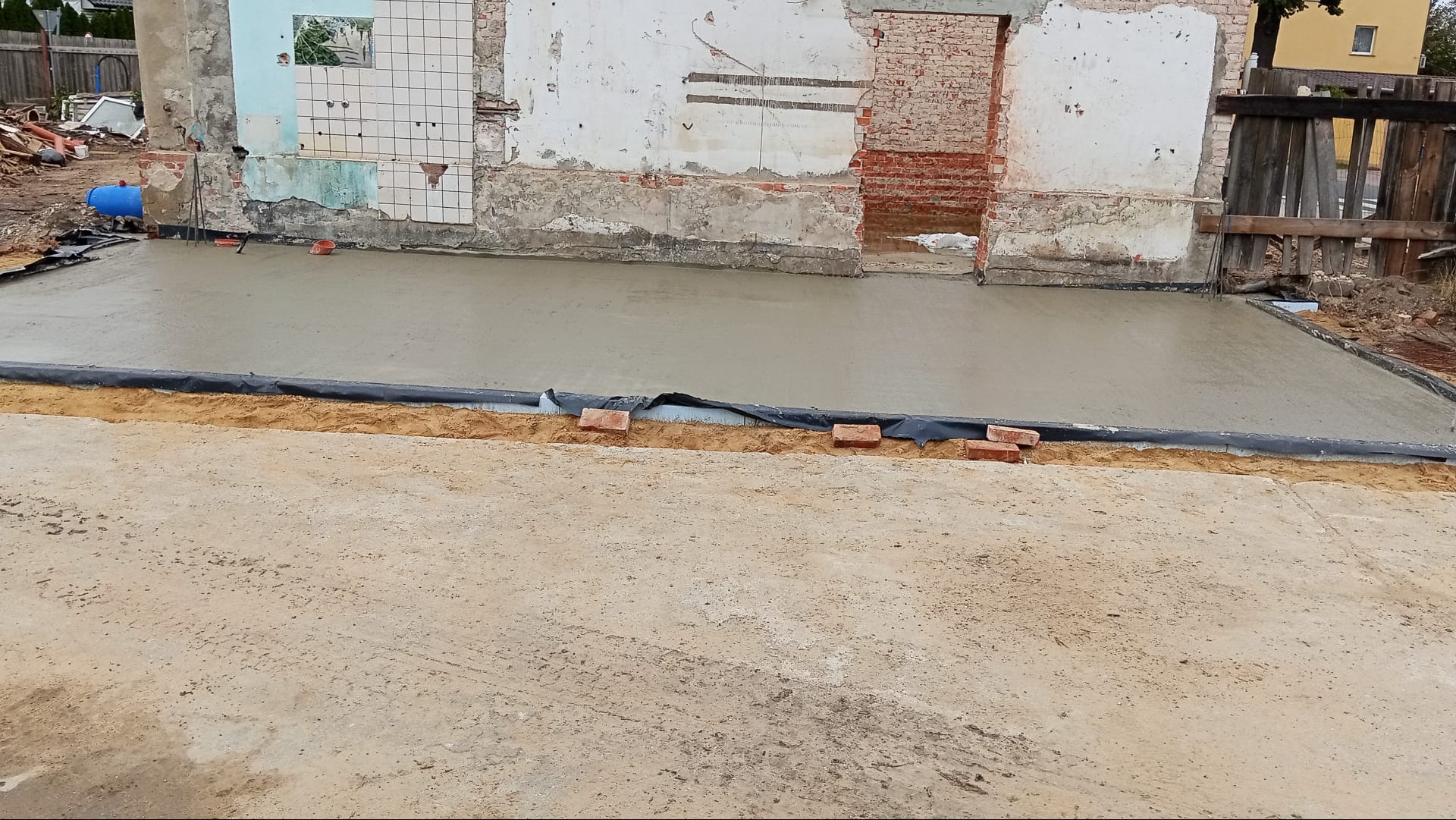 Wylany fundament pod budynek z widocznymi pozostałościami starej konstrukcji. Cegły oddzielają wylewkę od piasku. Fragmenty ścian z cegły i płytek.