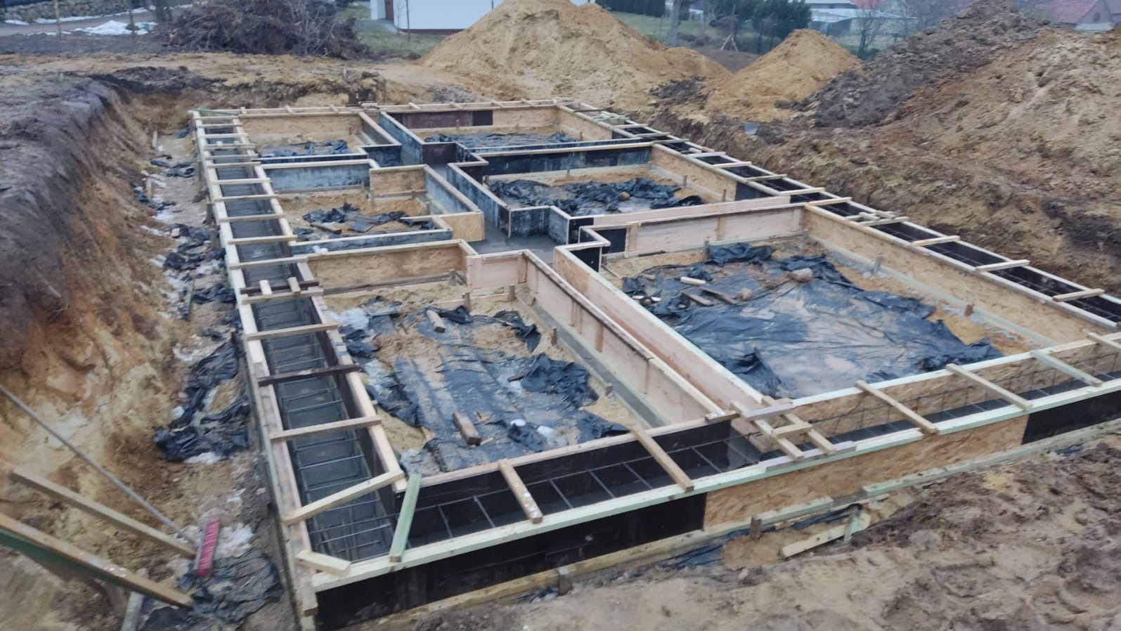 Szalunek fundamentów domu jednorodzinnego z widocznym zbrojeniem i folią ochronną, przygotowany do zalania betonem. W tle hałda ziemi.