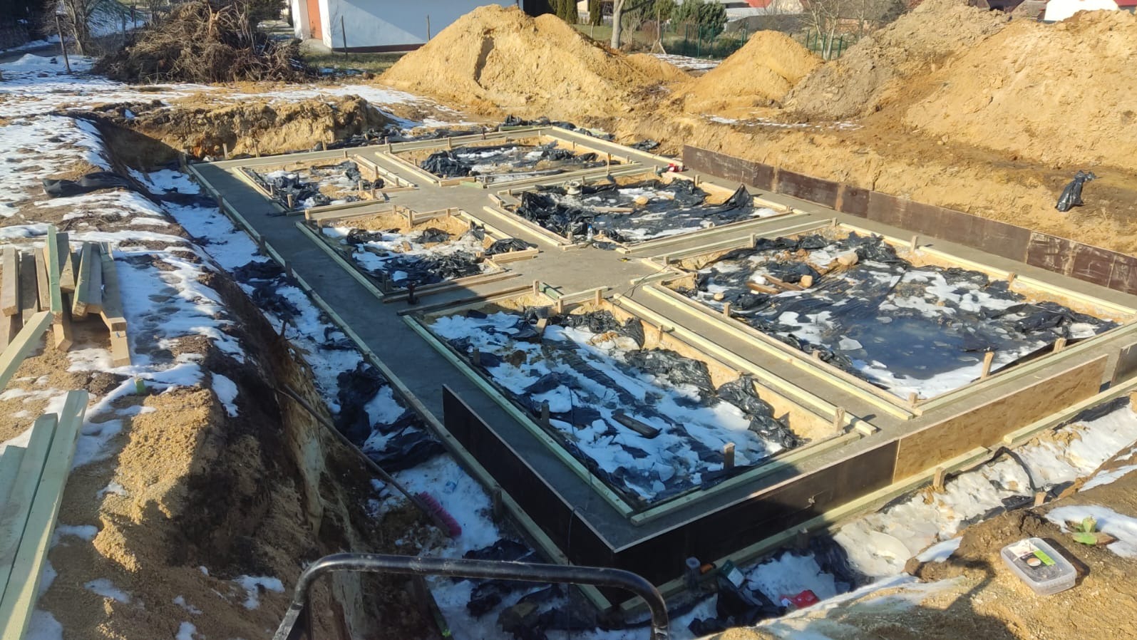 Zalane fundamenty domu jednorodzinnego z izolacją, widoczne szalunki i wykop, w tle hałdy ziemi. Zimowa sceneria z resztkami śniegu.