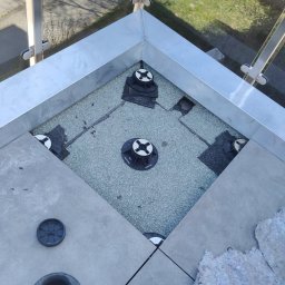 Rafał Stefański - Montaż podestu tarasowego z regulowanymi wspornikami i płytami betonowymi. Widok z góry na fragment tarasu z balustradą i elementami montażowymi.