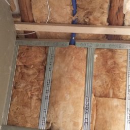 Rafał Stefański - Ocieplenie ściany wełną mineralną, widoczne profile metalowe, instalacja wodna z niebieskimi rurami i element podtynkowy baterii w wycięciu płyty gipsowej.