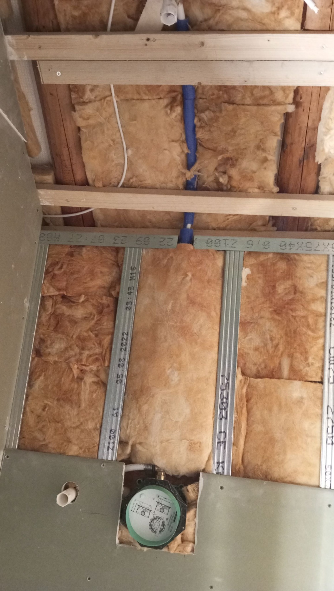 Ocieplenie ściany wełną mineralną, widoczne profile metalowe, instalacja wodna z niebieskimi rurami i element podtynkowy baterii w wycięciu płyty gipsowej.