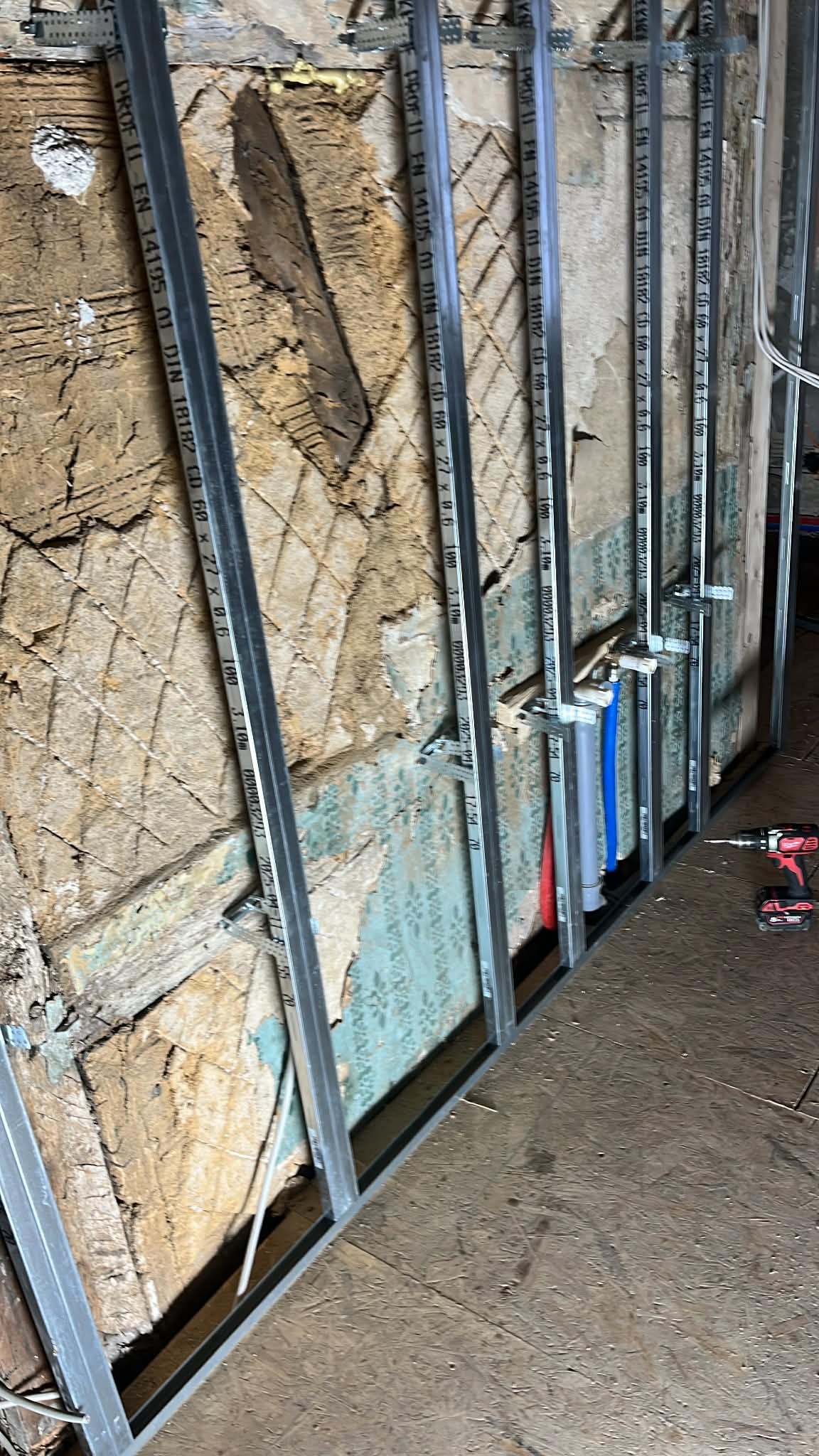 Metalowy stelaż przy starej ścianie z fragmentami tapety, widoczne rury instalacji wodnej i wiertarka. Etap prac przygotowawczych do wykończenia wnętrza.