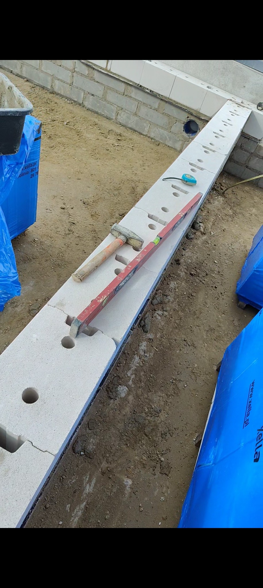 Wyrównany bloczek fundamentowy z otworami, poziomica i młotek na nim. W tle mur z bloczków i zapakowane bloczki. Widok z góry, lekki kąt.