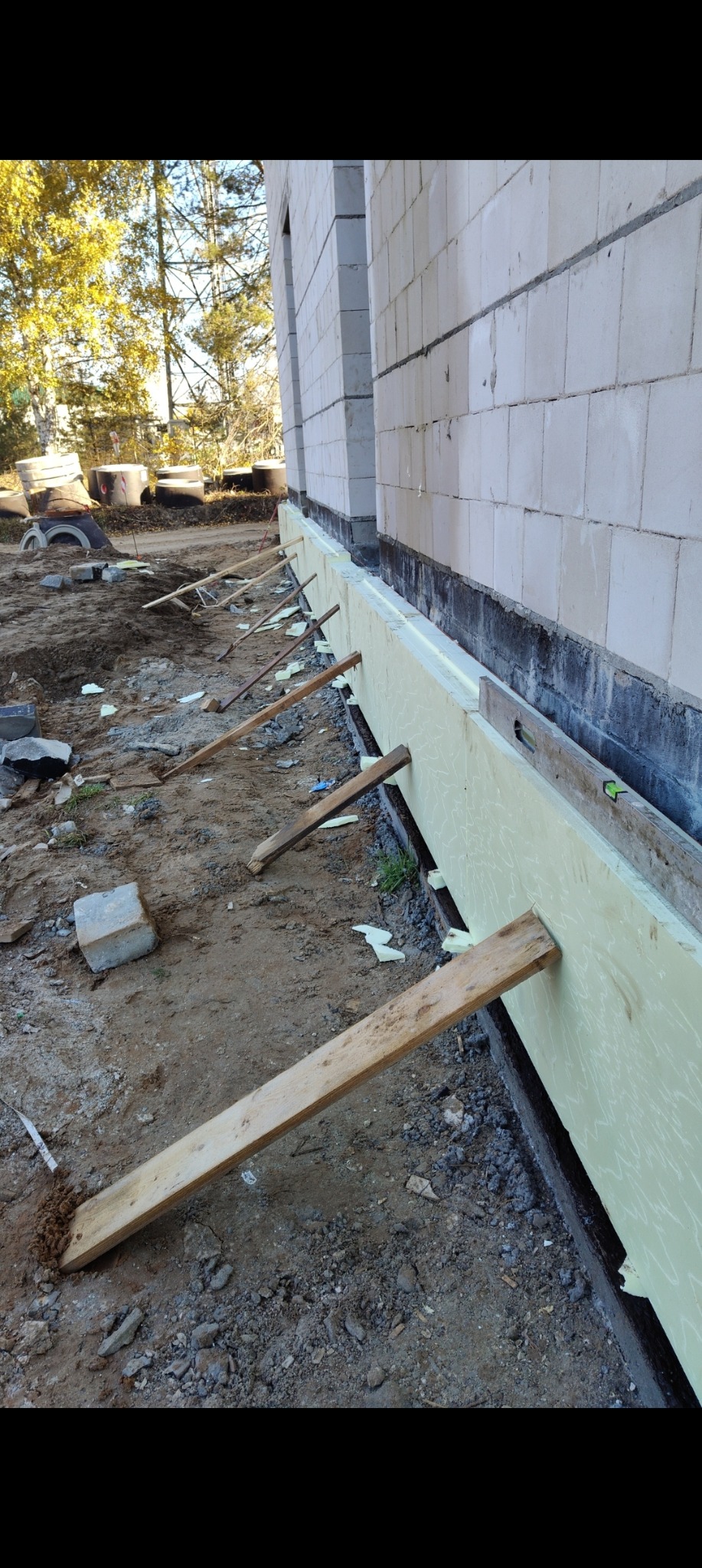 Ocieplenie fundamentów budynku z białego bloczka, zabezpieczone deskami. Widoczne elementy izolacji i poziomica. Prace ziemne w toku, teren budowy.