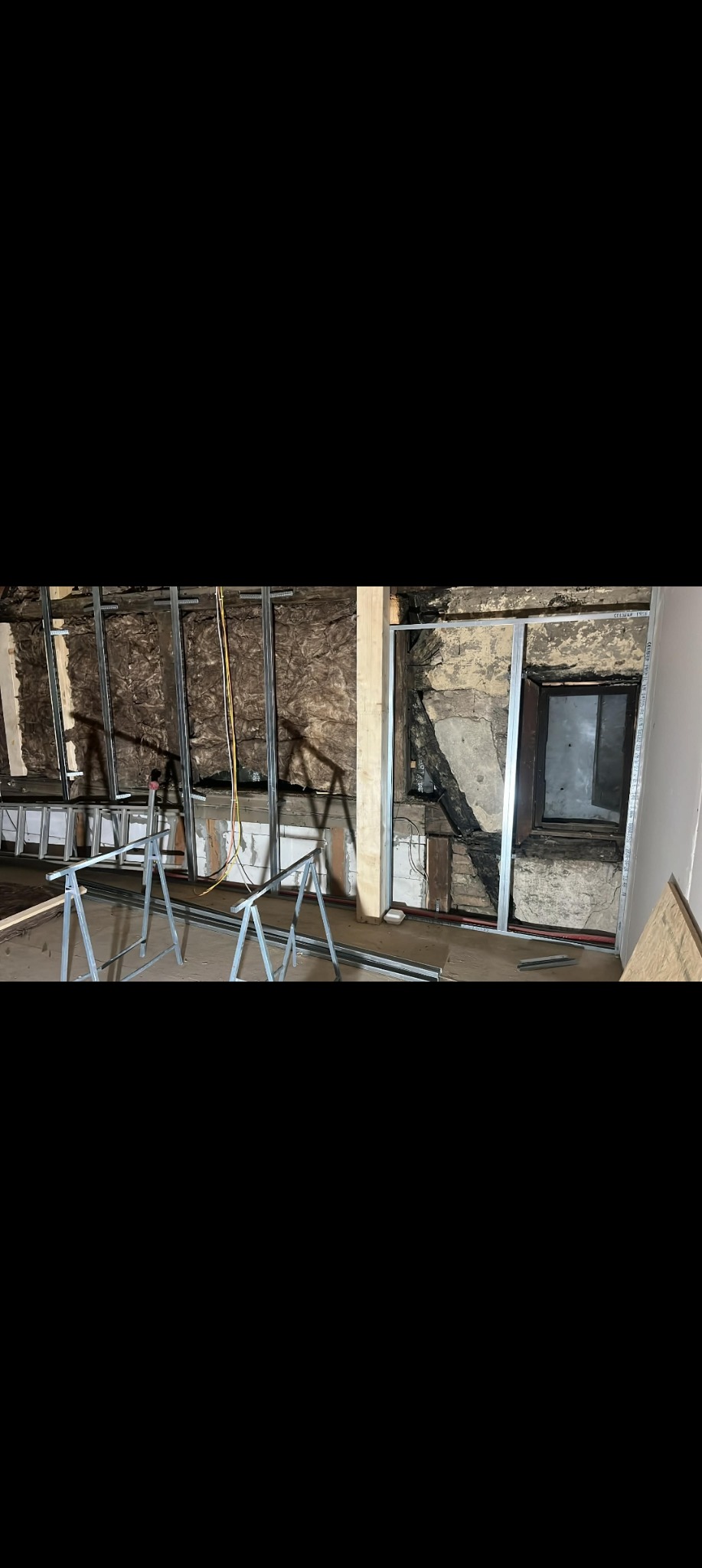 Remont starego domu: montaż metalowych profili na ścianach z widoczną izolacją i fragmentami oryginalnej konstrukcji. Widoczne rusztowania i okno.