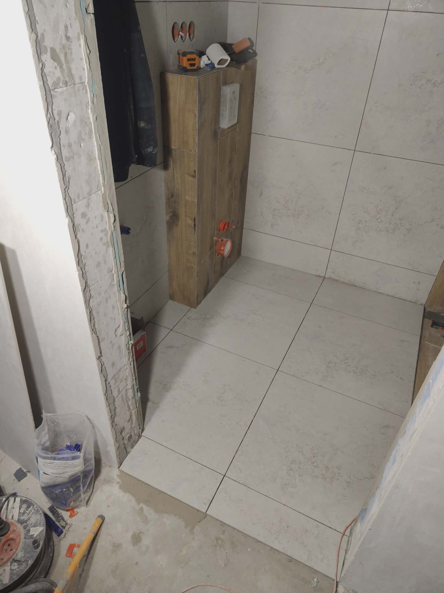 Układanie płytek w łazience: jasne, matowe płytki na podłodze i ścianach, zabudowa WC z drewnopodobną okleiną. Widoczne narzędzia glazurnicze i surowy stan wykończenia.