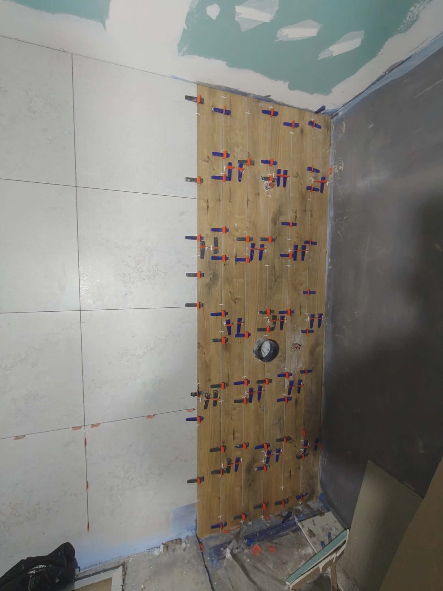 Układanie płytek imitujących drewno na ścianie w łazience, z użyciem klipsów poziomujących. Część ściany wykończona jasnymi płytkami, sufit w trakcie prac.