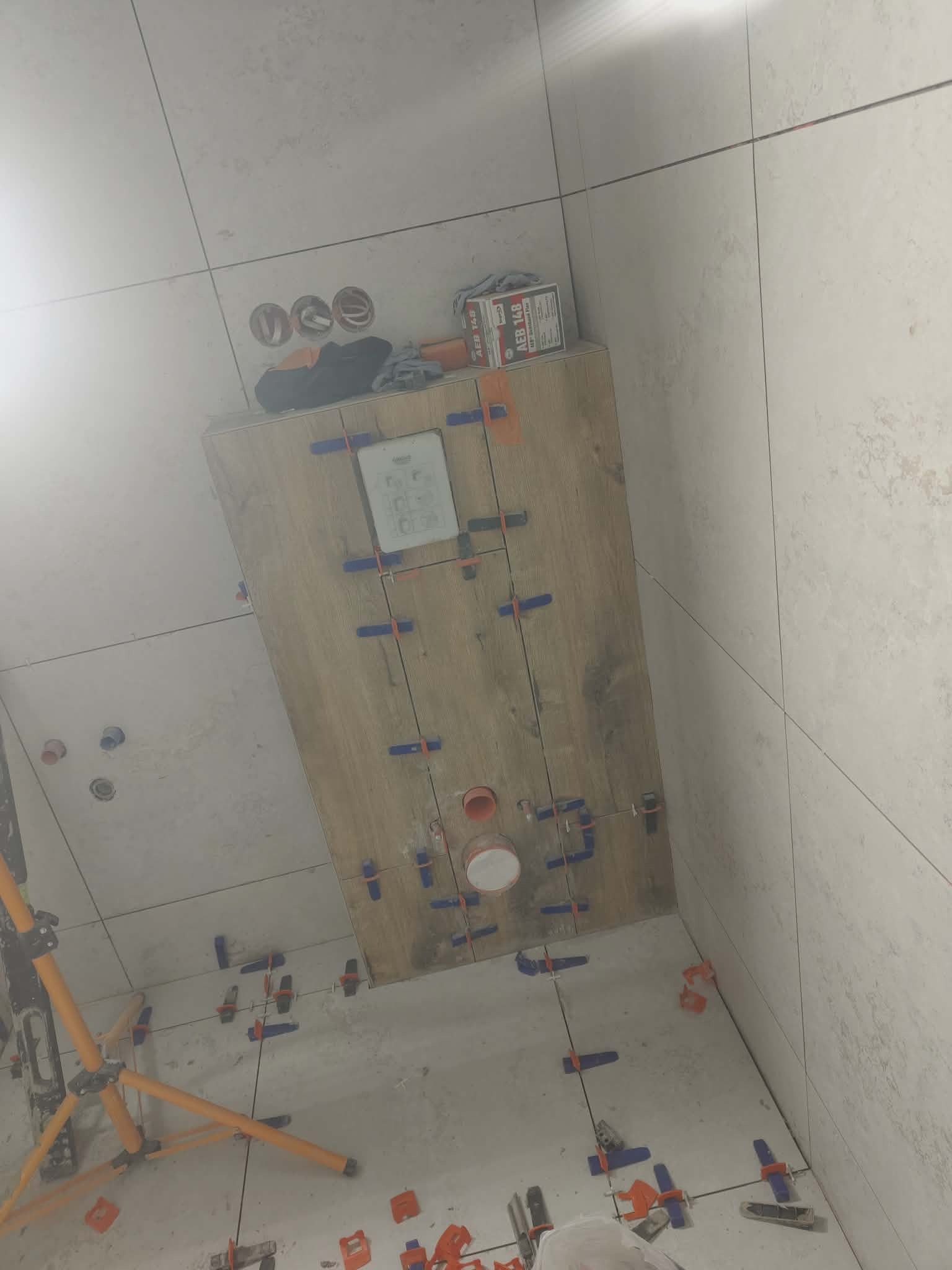 Układanie płytek imitujących drewno na podłodze w łazience, z użyciem klipsów poziomujących. Widoczne narzędzia glazurnicze i puszki instalacyjne w ścianie.