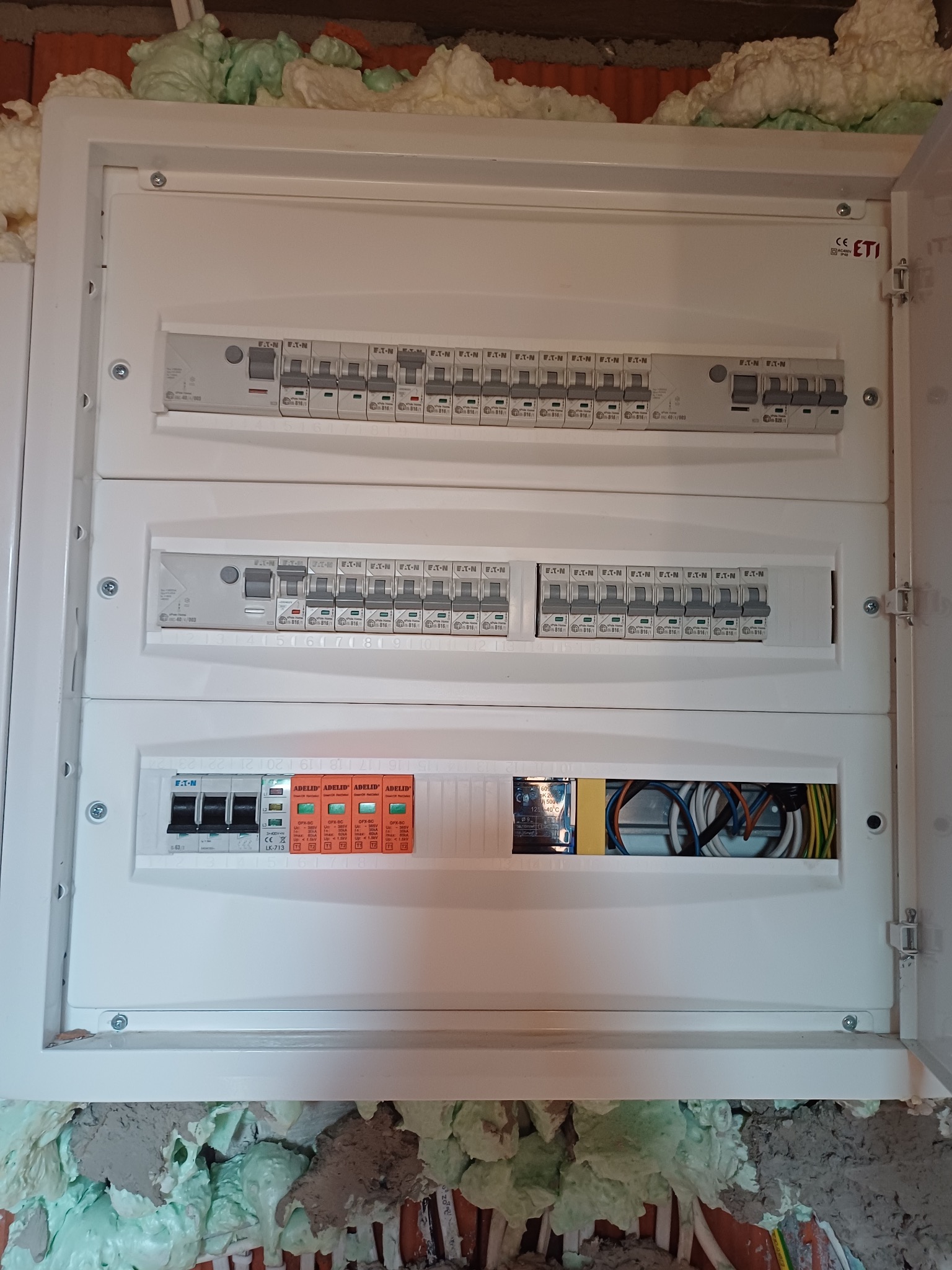 Rozdzielnica elektryczna w białej obudowie, widoczne zabezpieczenia i przewody. Montaż w trakcie prac wykończeniowych, pianka montażowa wokół obudowy.