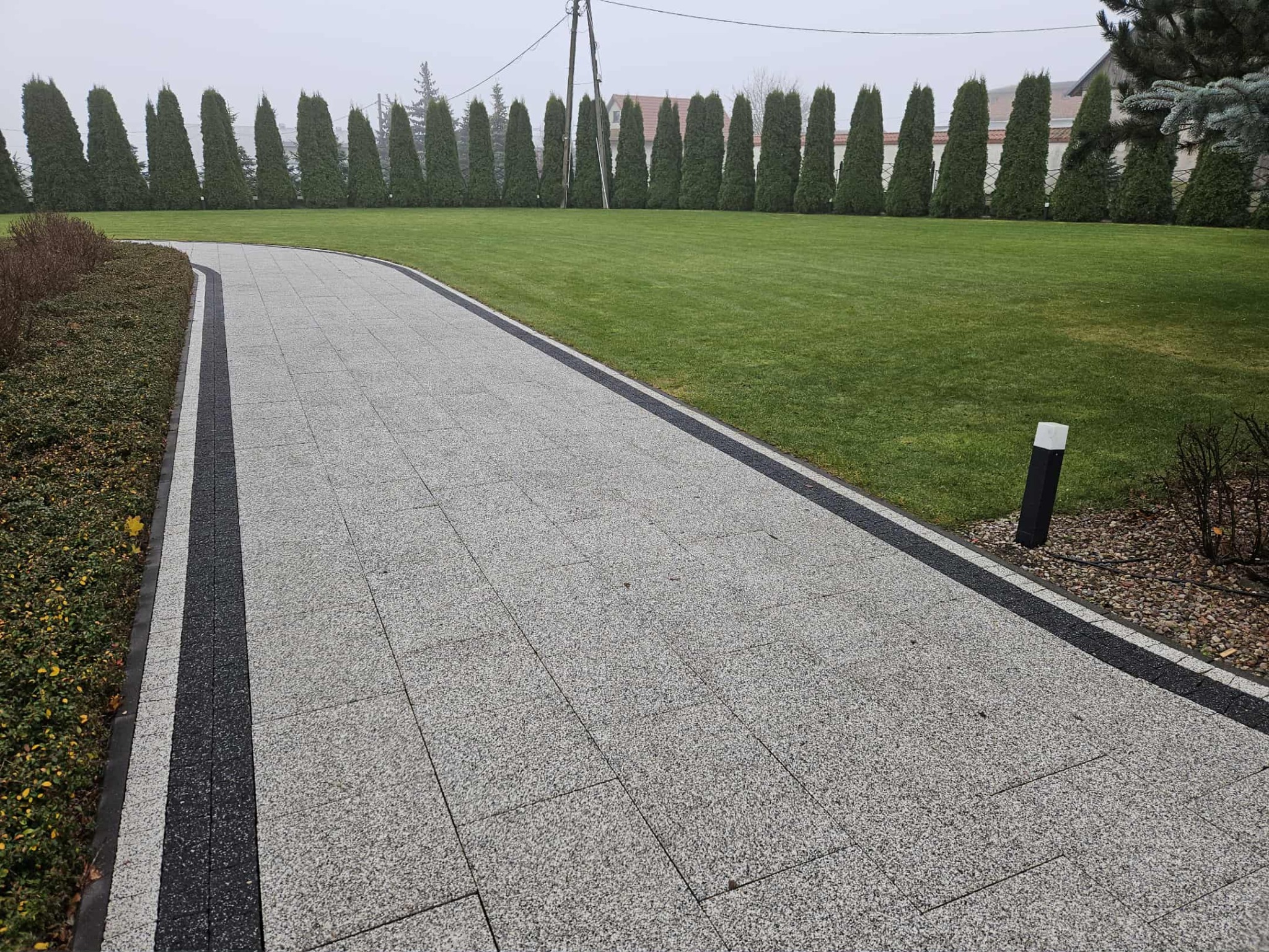 Kaskadowa ścieżka z jasnoszarej kostki brukowej z czarnymi obrzeżami, prowadząca przez trawnik z rzędem strzyżonych tuj w tle, oświetlona lampą ogrodową.