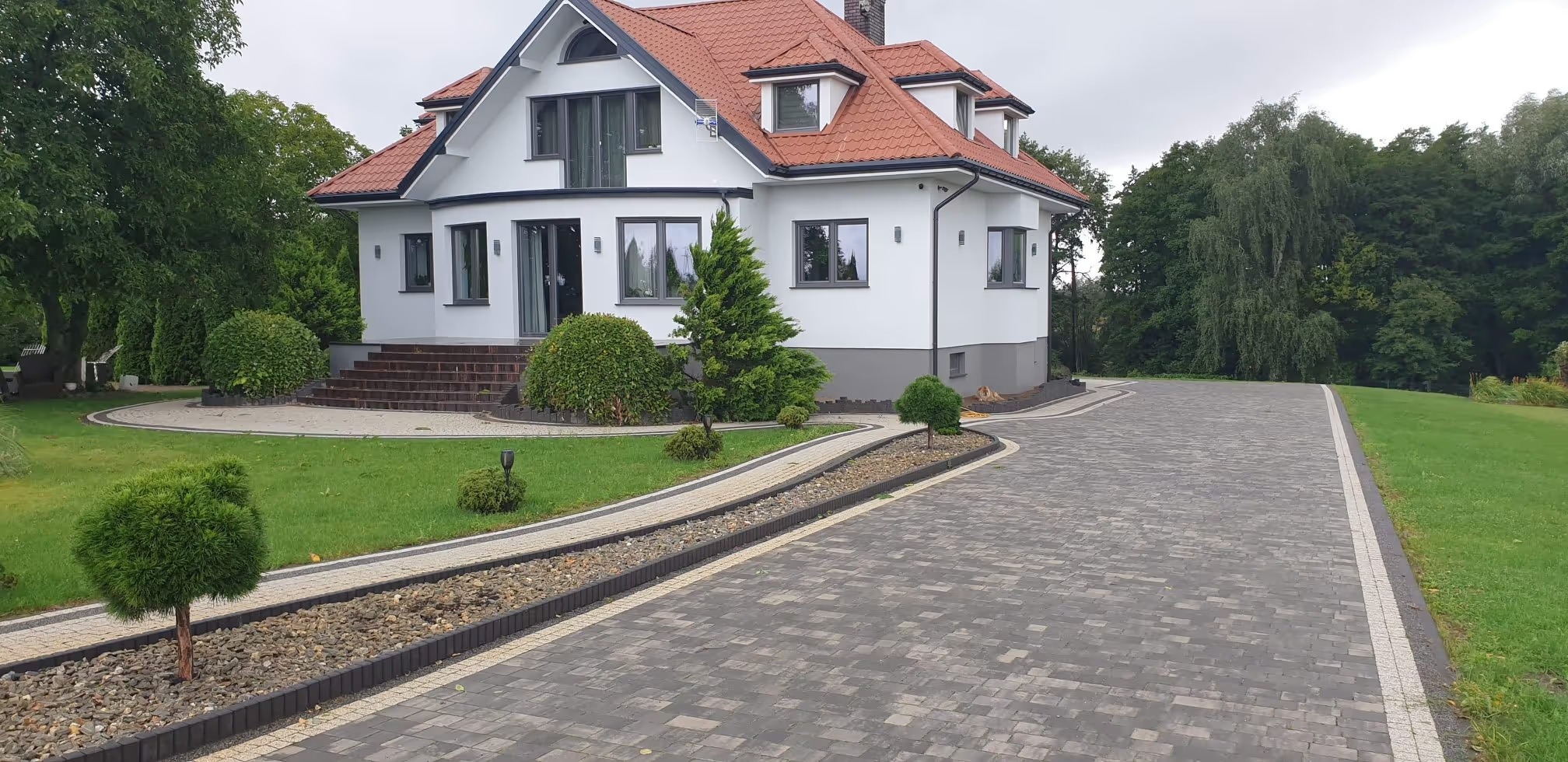 Elegancki podjazd z kostki brukowej prowadzący do nowoczesnego domu z czerwoną dachówką. Widoczne elementy drenażu i starannie utrzymany trawnik.
