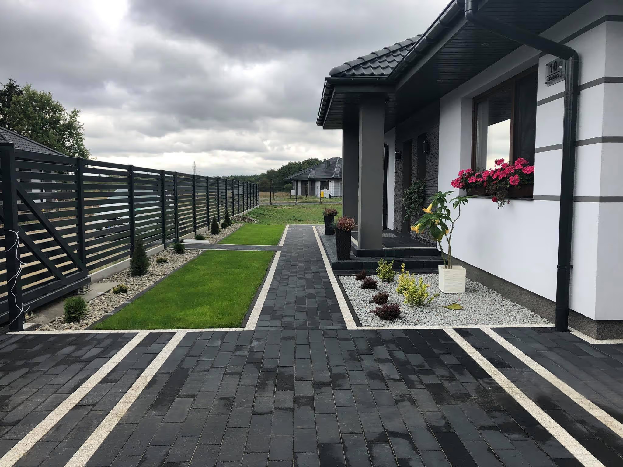 Nowoczesna posesja z czarną kostką brukową, białymi pasami i minimalistycznym ogrodem. Dom w tle z kwiatami w oknie. Całość utrzymana w stonowanej kolorystyce.