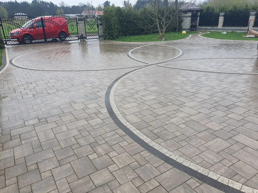 Ułożona kostka brukowa na podjeździe z dekoracyjnymi obramowaniami. W tle brama wjazdowa i czerwony samochód dostawczy. Widok na posesję w pochmurny dzień.