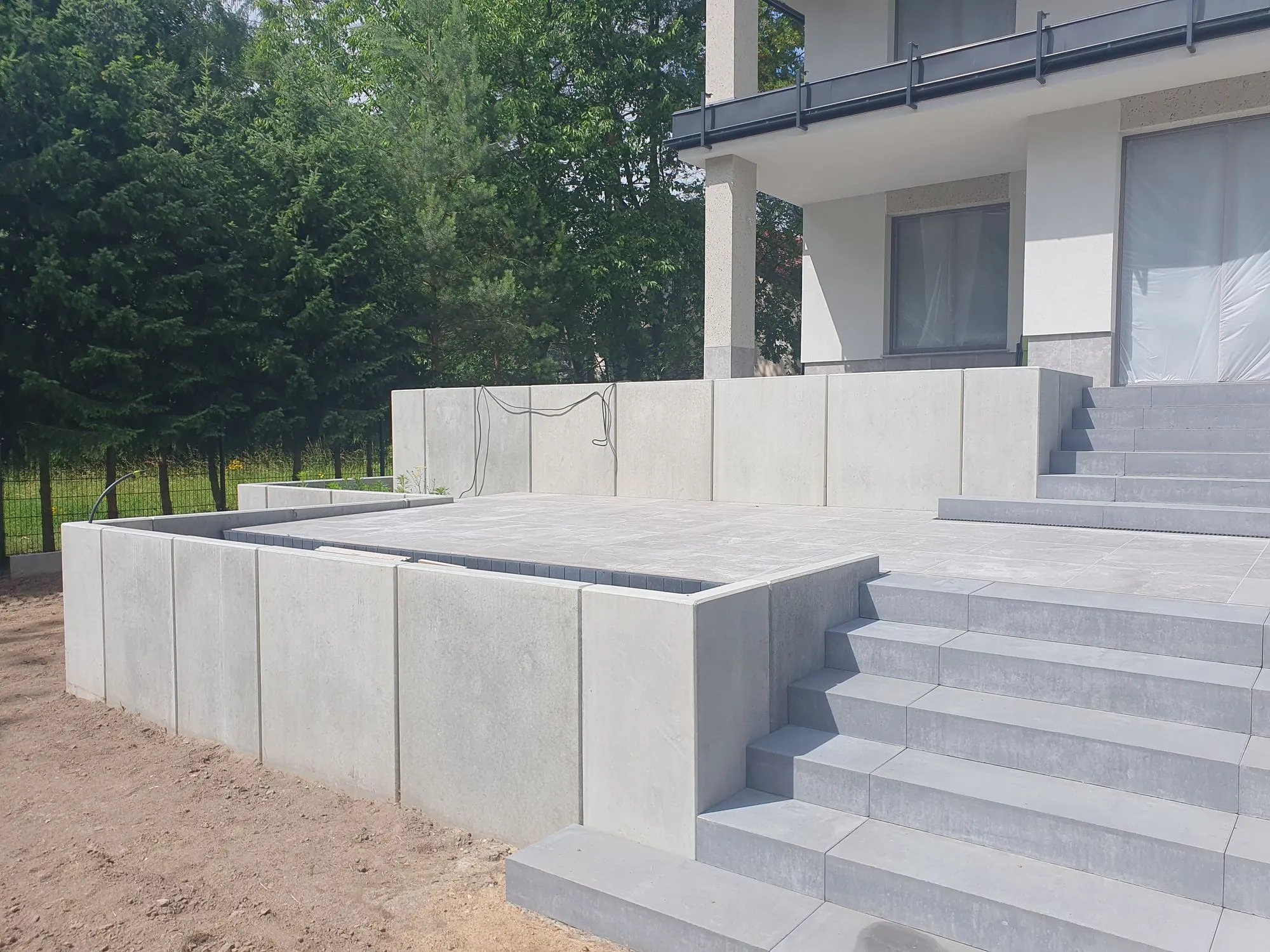 Nowoczesny taras z szarej kostki brukowej, schody prowadzące do domu, minimalistyczny design, widoczne połączenia kostki, surowe wykończenie, zieleń w tle.