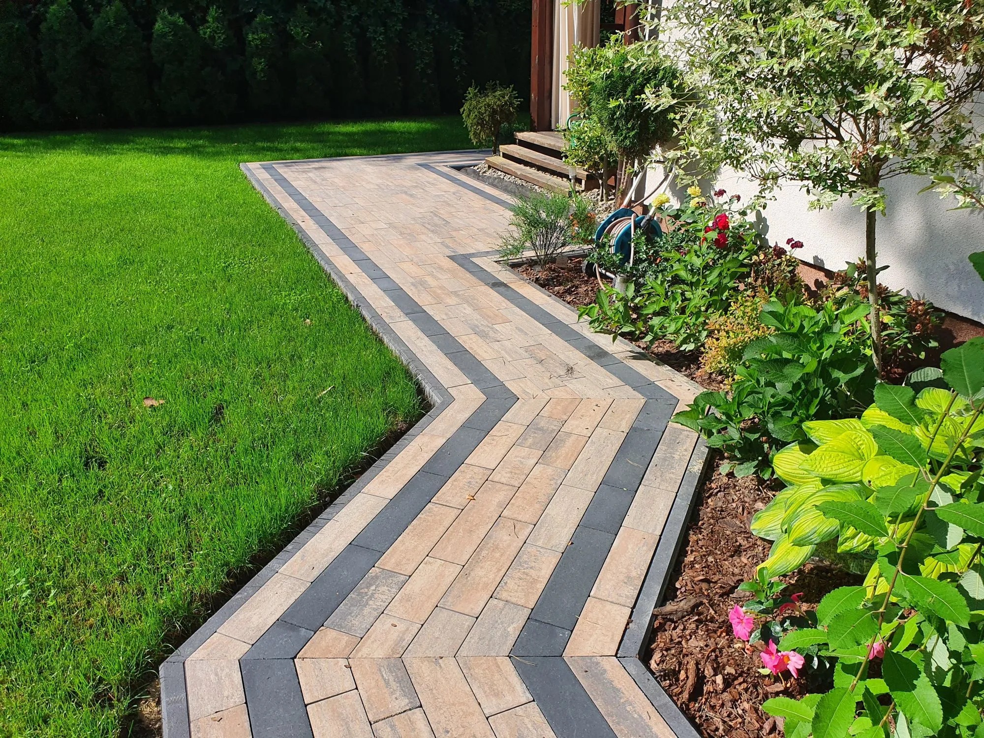 Kaskadowa ścieżka z kostki brukowej w odcieniach beżu i szarości, wijąca się przez trawnik i rabaty kwiatowe, prowadząca do wejścia domu z drewnianymi schodami.