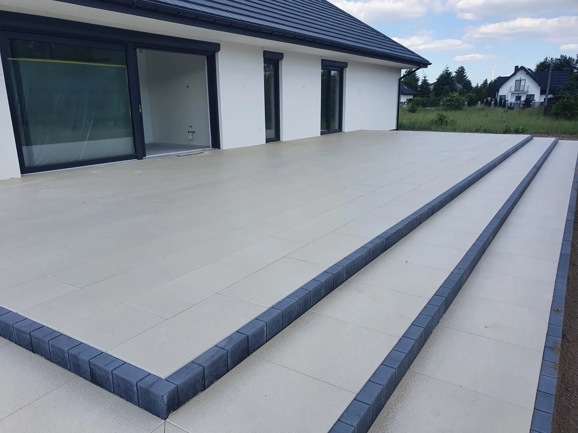 Jasna, wyłożona płytami tarasowymi przestrzeń przed domem z ciemną dachówką i obramowaniem. Schody z obrzeżami z kostki. Nowoczesny design.