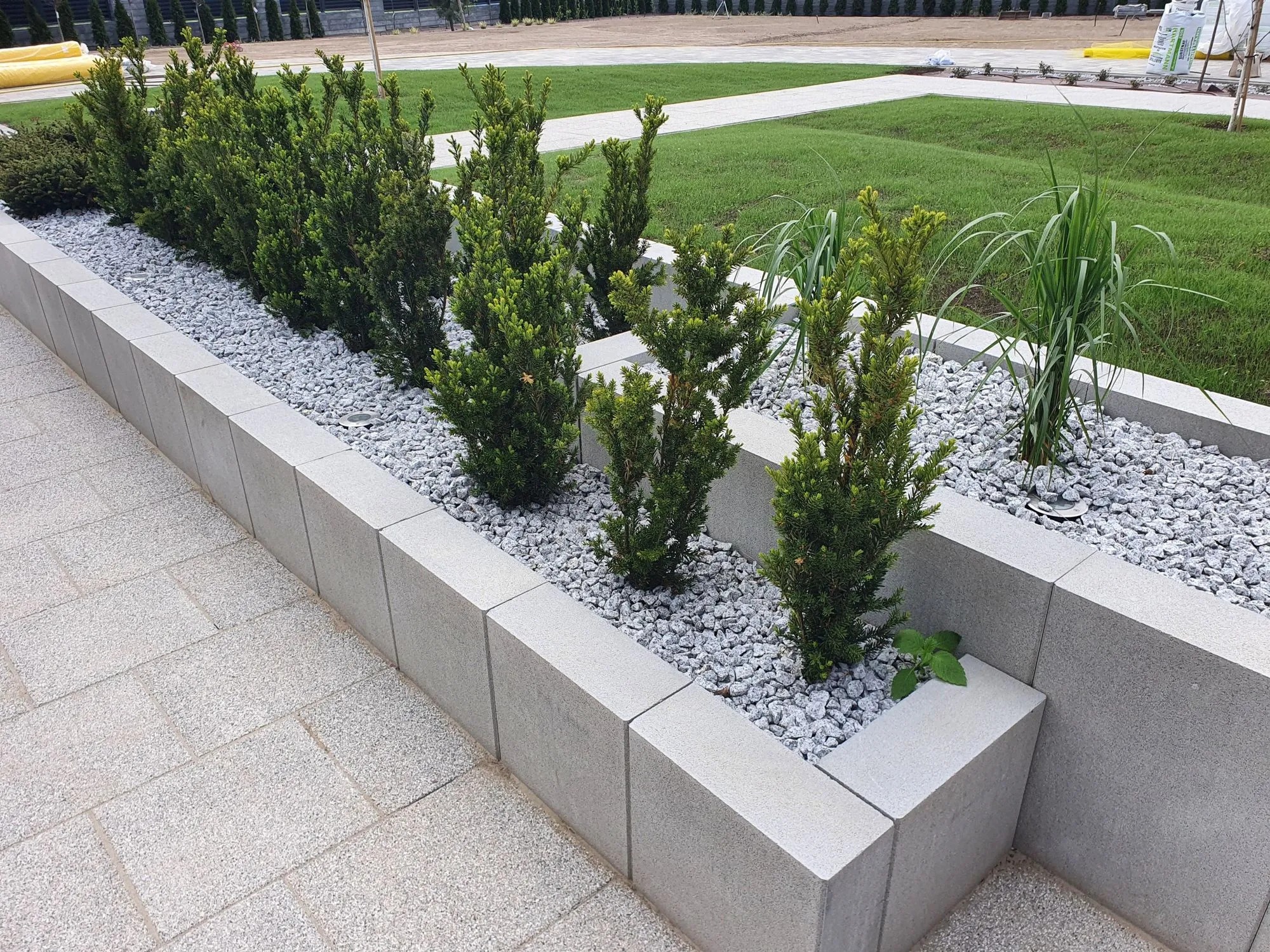 Kaskadowa rabata z szarej kostki, wypełniona białym grysem i zielonymi roślinami. Trawa i kostka brukowa w tle. Nowoczesny ogród.