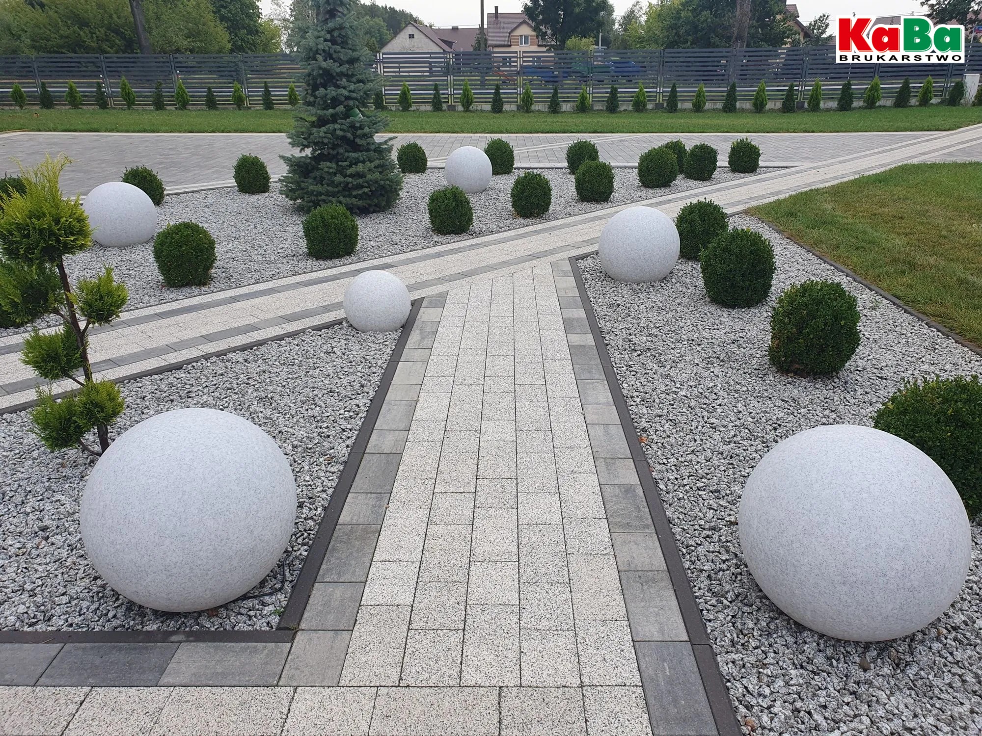 Ogród z ścieżką z kostki brukowej, ozdobnymi kulami i krzewami. Szara kostka, białe kule, zielone krzewy. Widok na zadbany teren z elementami dekoracyjnymi.