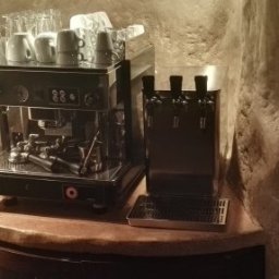 VENTONI SPÓŁKA Z OGRANICZONĄ ODPOWIEDZIALNOŚCIĄ - Profesjonalny ekspres do kawy z kranami do wody na blacie o fakturze kamienia, otoczony ścianami z surowego kamienia. Widoczne filiżanki i szklanki.