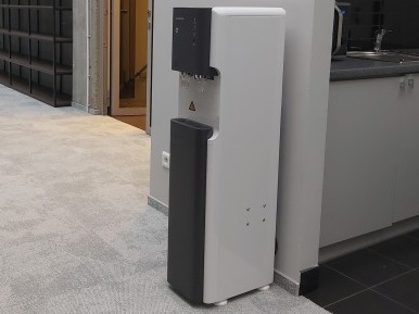 Nowoczesny dystrybutor wody w biurowym wnętrzu. Biało-czarna obudowa, widoczne krople wody. Minimalistyczny design, szara wykładzina podłogowa.