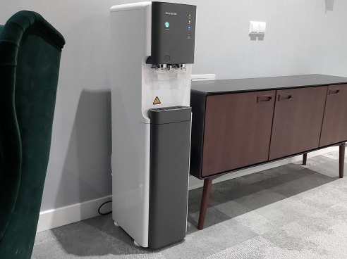 Nowoczesny dystrybutor wody w biurze, obok zielonego fotela i brązowej komody. Minimalistyczny design, szaro-biała kolorystyka, oświetlenie LED.