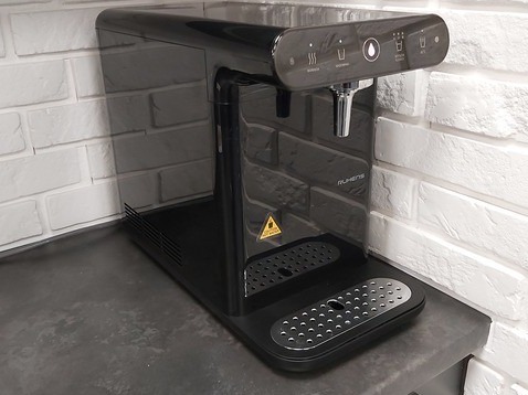 Czarny, nowoczesny dystrybutor wody marki Ruhens na blacie, z białym ceglanym tłem. Widoczne przyciski dotykowe i metalowa wylewka. Minimalistyczny design.