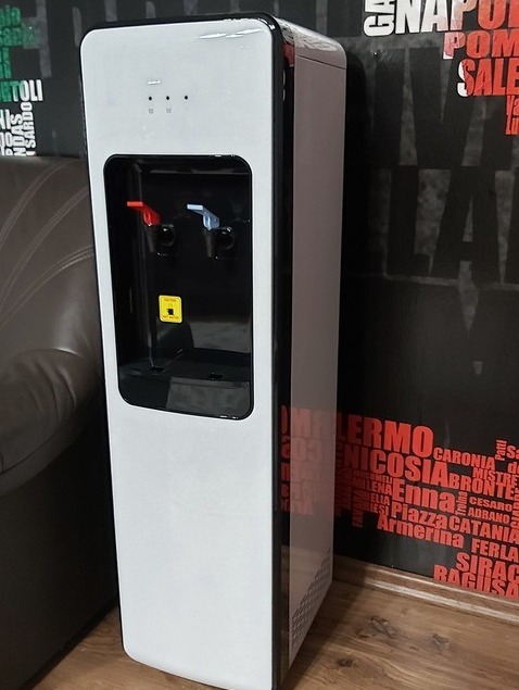 Elegancki, wolnostojący dystrybutor wody w kolorze białym z czarnymi akcentami, widoczny na tle ściany z włoskimi napisami. Nowoczesny design, idealny do biura.