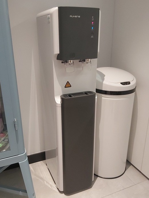 Nowoczesny dystrybutor wody Ruhens w biurowym wnętrzu, obok biały kosz na śmieci. Minimalistyczny design, oświetlenie LED, widoczne detale wykończenia.
