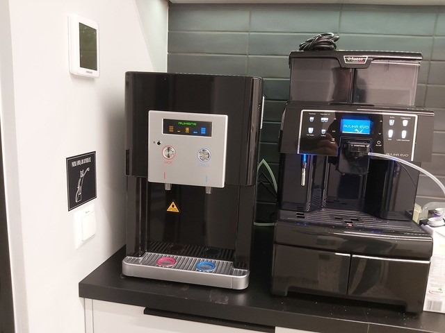 Czarny dystrybutor wody Mulhens obok ekspresu Saeco Aulika Evo na blacie. Dystrybutor z przyciskami ciepłej i zimnej wody, ekspres z wyświetlaczem i pojemnikiem na kawę.