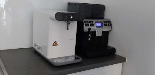 Nowoczesny dystrybutor wody obok ekspresu do kawy na blacie. Dystrybutor biały, ekspres czarny. Minimalistyczne wnętrze, jasne tło. Widoczne detale urządzeń.