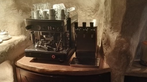 Profesjonalny ekspres do kawy z kranami do wody na blacie o fakturze kamienia, otoczony ścianami z surowego kamienia. Widoczne filiżanki i szklanki.