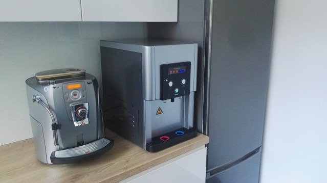 Srebrny ekspres do kawy i dystrybutor wody obok lodówki w jasnej kuchni. Dystrybutor ma panel sterowania z wyświetlaczem i przyciskami.