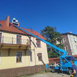 Zaczarowany ogród - Praca na wysokościach w Gniewino: Błękitny podnośnik koszowy przy budynku z czerwoną dachówką. Osoba w koszu na dachu. Usługi wysokościowe.