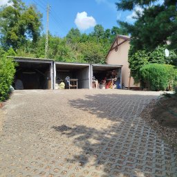 Zaczarowany ogród - Ułożona szara kostka ażurowa na podjeździe, z widocznymi trzema garażami blaszanymi w tle, otoczona zielenią i drzewami w słoneczny dzień.