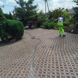 Zaczarowany ogród - Czyszczenie kostki ażurowej wodą i szczotką przez pracownika w jaskrawozielonym kombinezonie. Wąż ogrodowy na pierwszym planie. Tło: zieleń ogrodu.