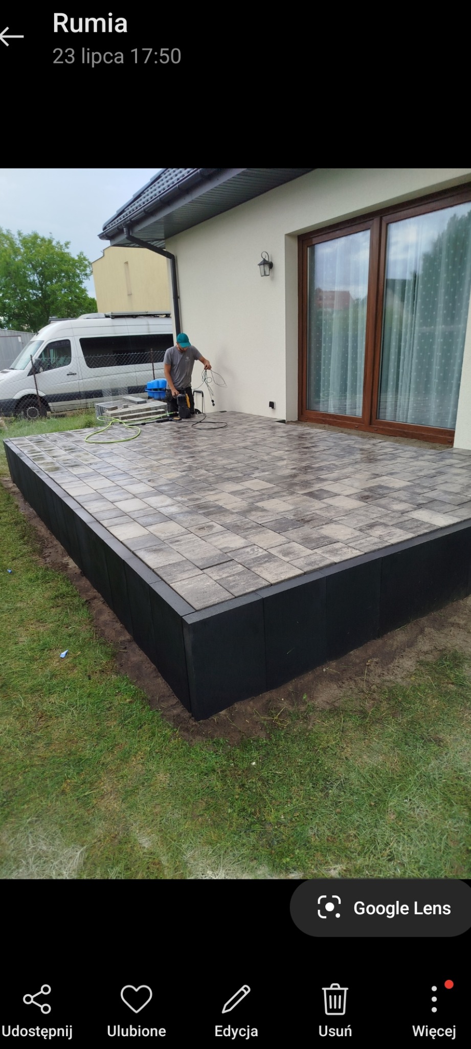 Ułożona kostka brukowa na podwyższonym tarasie przy domu. Pracownik czyści powierzchnię myjką ciśnieniową. Widoczny fragment elewacji i okna.