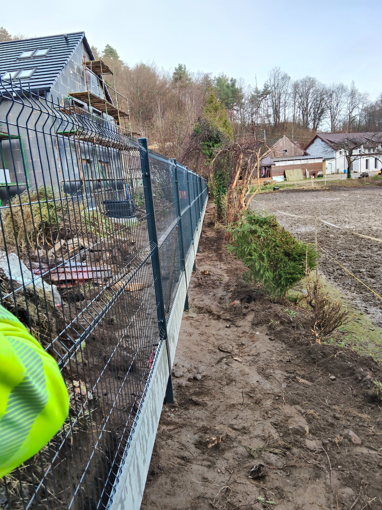 Nowoczesne, panelowe ogrodzenie z podmurówką betonową w trakcie montażu. Widoczny wykop pod fundament, teren po niwelacji i fragment budynku w tle.