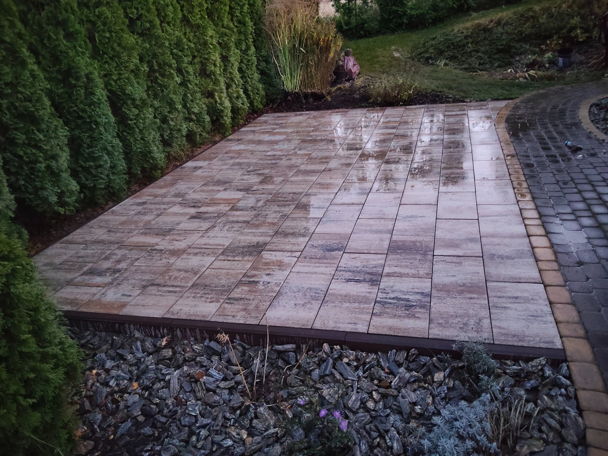 Ułożona, prostokątna kostka brukowa w odcieniach beżu i brązu, obramowana ciemnymi kamieniami i zielonymi tujami. Widoczny fragment ścieżki z kostki po prawej stronie.