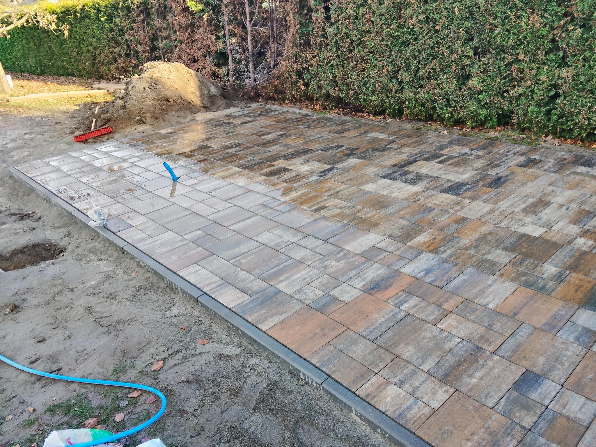 Układanie kostki brukowej: Częściowo ułożona, wielobarwna kostka brukowa z obrzeżami na podsypce piaskowej, widoczny stos piasku i narzędzia.